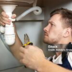 young handsome man plumber in uniform fixing the sink with pliers in his hand sitting on the kitchen floor professional plumbing repair service.